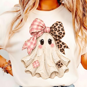 Puede incluir: Una camiseta color crema con el gráfico de un fantasma blanco de ojos negros grandes y mejillas rosadas. El fantasma lleva un lazo grande con un lado de patrón de cuadros vichy rosa y el otro con estampado de leopardo marrón y parches.