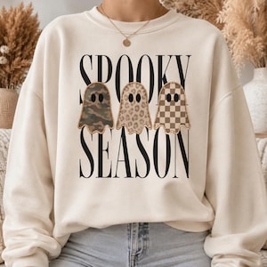 May include: A cream-colored crewneck sweatshirt featuring the black text SPOOKY SEASON. Centered between the words are three ghost illustrations, each with a different pattern: one in green camouflage, one in leopard print, and one in a brown checkerboard.