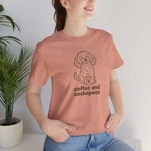 Peut inclure: T-shirt pêche avec un dessin au trait d'un chien Cockapoo tenant une tasse de café. Le texte "coffee and cockapoos" est imprimé en dessous. Le t-shirt est fait d'une matière douce.