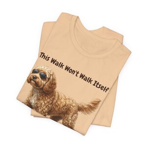 Op de afbeelding: Een lichtperzikkleurig T-shirt met een cartoonhond met een zonnebril. De hond is lichtbruin met krullend haar. De tekst "This Walk Won't Walk Itself" staat in het zwart op de bovenkant van het shirt. Het shirt is opgevouwen.