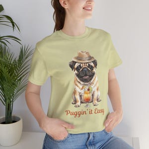 May include: Light yellow t-shirt featuring a watercolor illustration of a pug wearing a straw hat and holding a cocktail. The text "Puggin' it Easy" is printed below the pug. The shirt is made of soft material.