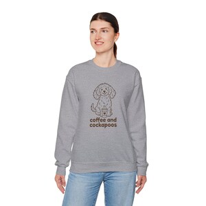 Op de afbeelding: Een grijze sweatshirt met een bruine illustratie van een Cockapoo-hond die een koffiekopje vasthoudt. De tekst "coffee and cockapoos" staat eronder. De sweatshirt wordt gedragen door een persoon die een blauwe jeans draagt.