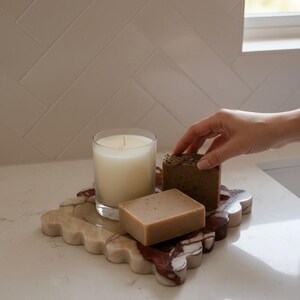 May include: A marble tray with a candle and two soaps. The tray has a scalloped edge and is beige and burgundy. The candle is in a clear glass, and the soaps are brown. The items are on a white surface.