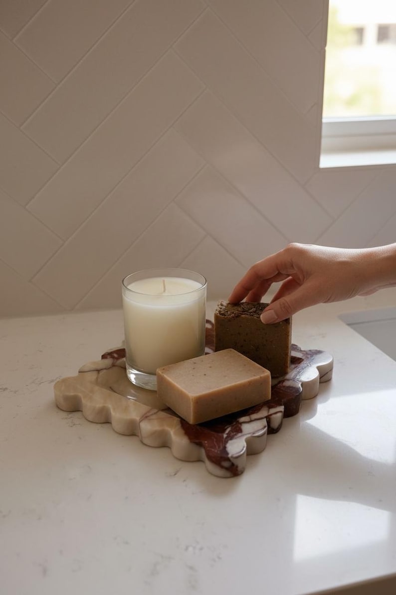 May include: A marble tray with a candle and two soaps. The tray has a scalloped edge and is beige and burgundy. The candle is in a clear glass, and the soaps are brown. The items are on a white surface.