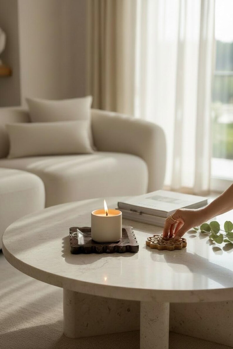 May include: A burning, cream-coloured candle in a cylindrical holder sits on a dark, textured coaster. A decorative wooden coaster is next to it on a round, light-coloured marble table. A sofa and books are in the background.