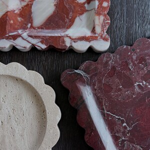 May include: Three decorative trays with scalloped edges. One tray is a light beige colour, another is a deep red with white veining, and the third is red with white and beige veining. The trays are made of stone and are arranged on a dark wood surface.