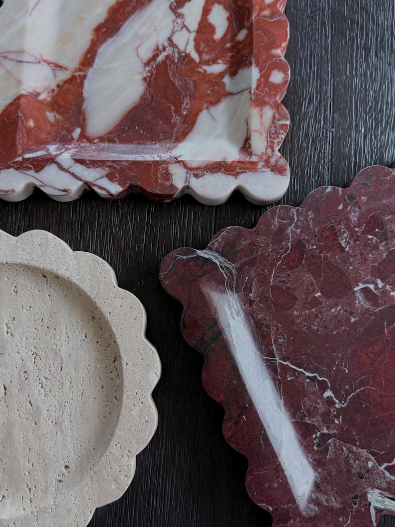 May include: Three decorative trays with scalloped edges. One tray is a light beige colour, another is a deep red with white veining, and the third is red with white and beige veining. The trays are made of stone and are arranged on a dark wood surface.