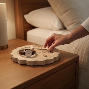 May include: A beige, scalloped-edge tray holds a brown leather-banded watch, gold-rimmed glasses, and a small gold ring. The tray sits on a wooden bedside table next to a bed with white bedding. A person's hand reaches for the glasses.