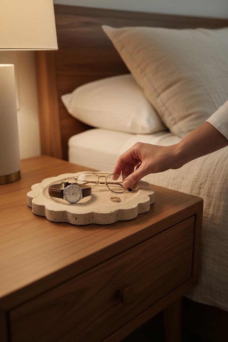 May include: A beige, scalloped-edge tray holds a brown leather-banded watch, gold-rimmed glasses, and a small gold ring. The tray sits on a wooden bedside table next to a bed with white bedding. A person's hand reaches for the glasses.