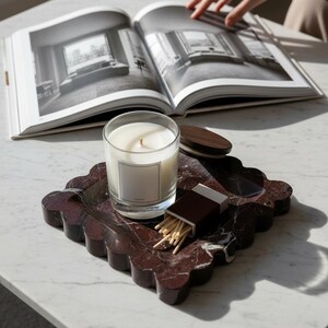 May include: A candle in a clear glass jar with a blank label sits on a decorative, scalloped, burgundy marble tray. A box of matches and a wooden lid are also on the tray. An open book with black and white photos is in the background.