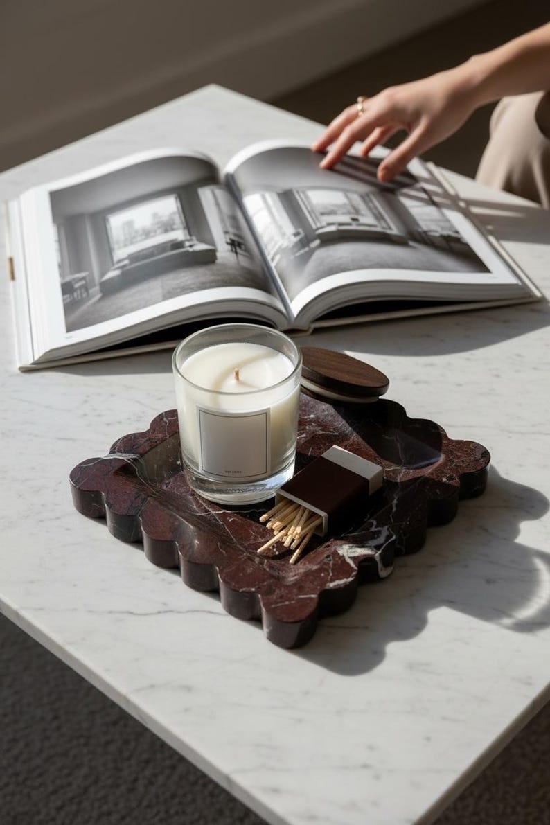 May include: A candle in a clear glass jar with a blank label sits on a decorative, scalloped, burgundy marble tray. A box of matches and a wooden lid are also on the tray. An open book with black and white photos is in the background.