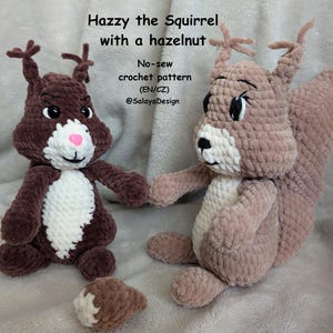 May include: Two plush squirrel toys made of soft yarn. One is dark brown with a white chest and pink nose, the other is light brown with a white chest. A small crocheted hazelnut sits between them. Text reads: Hazzy the Squirrel with a hazelnut, No-sew crochet pattern (EN/CZ), @SalayaDesign.