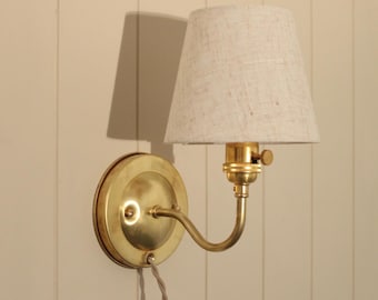 May include: A wall-mounted lamp featuring a circular brass backplate and a curved brass arm. The fixture includes a small, tapered, light beige fabric lampshade and a brass socket with a side-mounted turn knob switch, connected to a fabric cord.