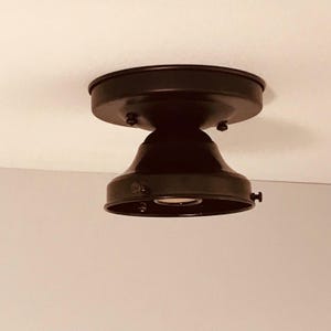 May include: A matte black flush mount ceiling light fixture featuring a circular base plate and a bell-shaped shade. The fixture includes small side screws for securing a glass component and a central socket for a single light bulb installation.