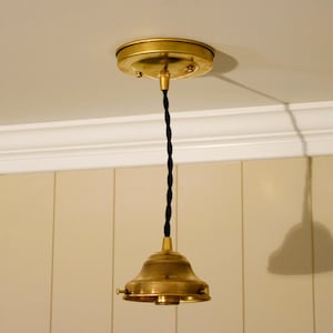 May include: A brass pendant light fixture featuring a circular ceiling canopy, a black twisted fabric cord, and a bell-shaped brass shade holder. The metal components have a warm, brushed finish, and the fixture hangs vertically from the ceiling.