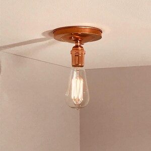May include: A copper-toned flush mount ceiling light fixture featuring a circular base and a single socket. A clear glass Edison-style light bulb with a visible vertical filament is screwed into the socket, emitting a warm, soft glow.