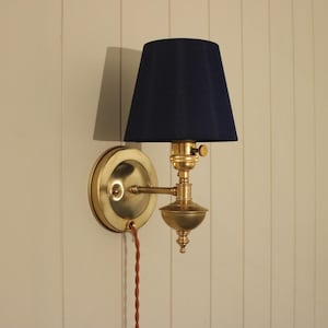 May include: A wall-mounted lamp featuring a polished brass circular base and a matching brass arm that supports a dark navy blue fabric lampshade. The fixture includes a small brass knob switch and a twisted brown electrical cord hanging downward.