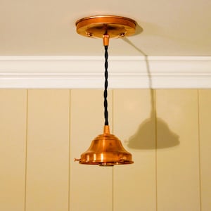 May include: A copper pendant light fixture featuring a circular ceiling canopy, a black twisted fabric cord, and a bell-shaped copper shade. The metal components have a polished finish, and the fixture is suspended vertically from the ceiling.