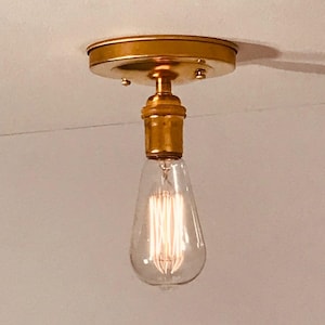 May include: A flush mount ceiling light fixture with a metallic gold finish circular base and a matching socket. The fixture holds a clear glass teardrop-shaped Edison light bulb with visible vertical glowing filaments inside the glass casing.