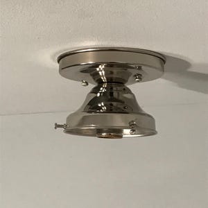 May include: A polished nickel flush mount ceiling light fixture base with a circular canopy, a bell-shaped housing, and three small side screws for securing a glass shade. The metal surface is reflective and smooth, showing a metallic silver finish.
