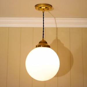 May include: A spherical white glass globe pendant light fixture suspended by a black twisted fabric cord from a circular brass ceiling canopy. The globe is secured to the cord with a brass metal cap and hardware, creating a clean lighting design.