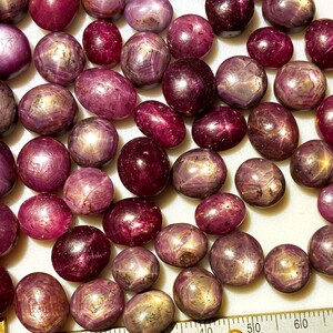 May include: A collection of 30+ loose, cabochon cut, star ruby gemstones. The stones are a deep reddish-purple color with a star-like pattern visible in the center of each stone.