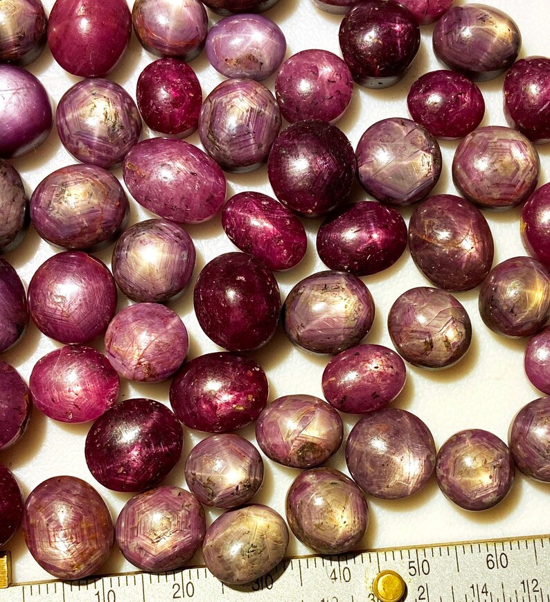 May include: A collection of 30+ loose, cabochon cut, star ruby gemstones. The stones are a deep reddish-purple color with a star-like pattern visible in the center of each stone.