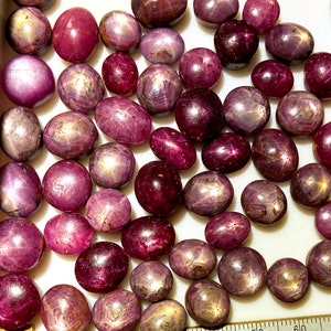 May include: A collection of pink and purple star ruby cabochons. The stones are smooth and polished, with a star-shaped pattern visible on their surface.
