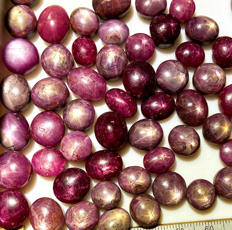 May include: A collection of pink and purple star ruby cabochons. The stones are smooth and polished, with a star-shaped pattern visible on their surface.