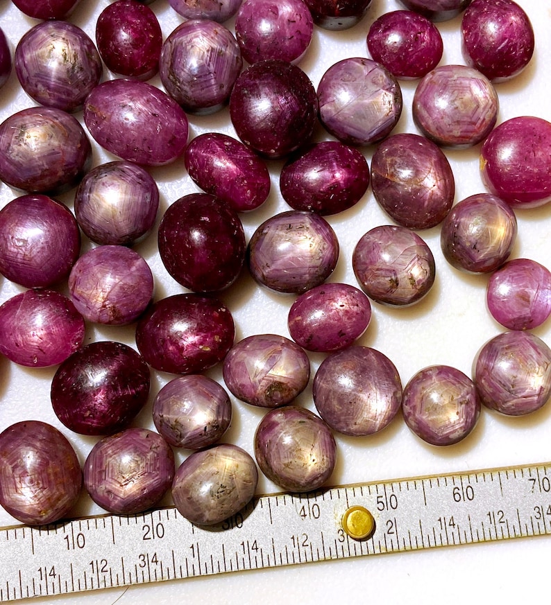May include: A collection of 30 oval-shaped, pink star ruby cabochons. The stones have a silvery sheen and are arranged on a white surface with a ruler for scale.