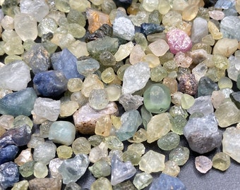 May include: A collection of small, rough, and faceted gemstones in various shades of blue, green, and yellow. The gemstones are scattered on a dark surface.