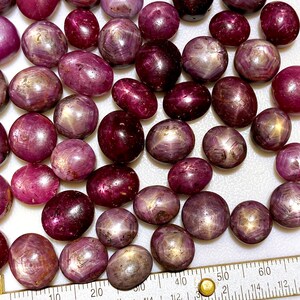 May include: A collection of 40+ star ruby cabochons in various shades of pink and red. The stones are oval shaped and have a star-like pattern in the center. The stones are arranged on a white surface with a ruler for scale.
