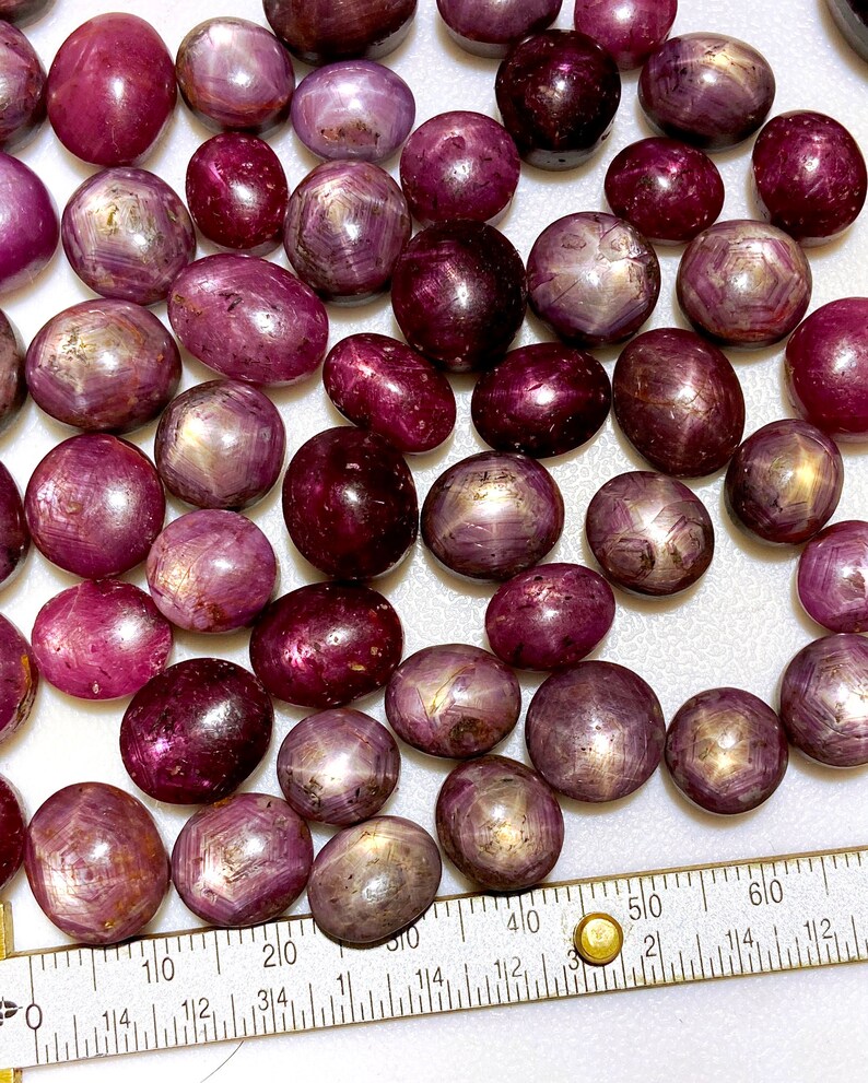 May include: A collection of 40+ star ruby cabochons in various shades of pink and red. The stones are oval shaped and have a star-like pattern in the center. The stones are arranged on a white surface with a ruler for scale.