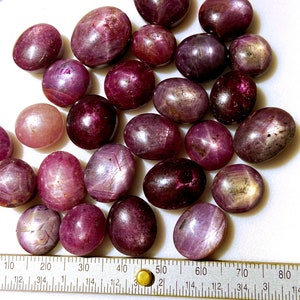 May include: A collection of 25 oval-shaped, star ruby cabochons in shades of pink and purple. The stones are arranged on a white surface with a ruler for scale.