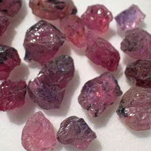 May include: A collection of raw, unpolished gemstones in various shades of pink, magenta, and deep purple. These small, irregular mineral fragments feature faceted-like textures and translucent surfaces, scattered across a plain white surface.