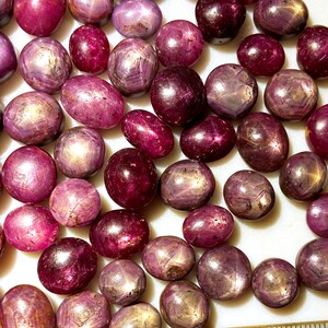 May include: A collection of pink star ruby cabochons, each with a six-pointed star in the center. The stones are various sizes and shapes, and they are all polished to a high shine.