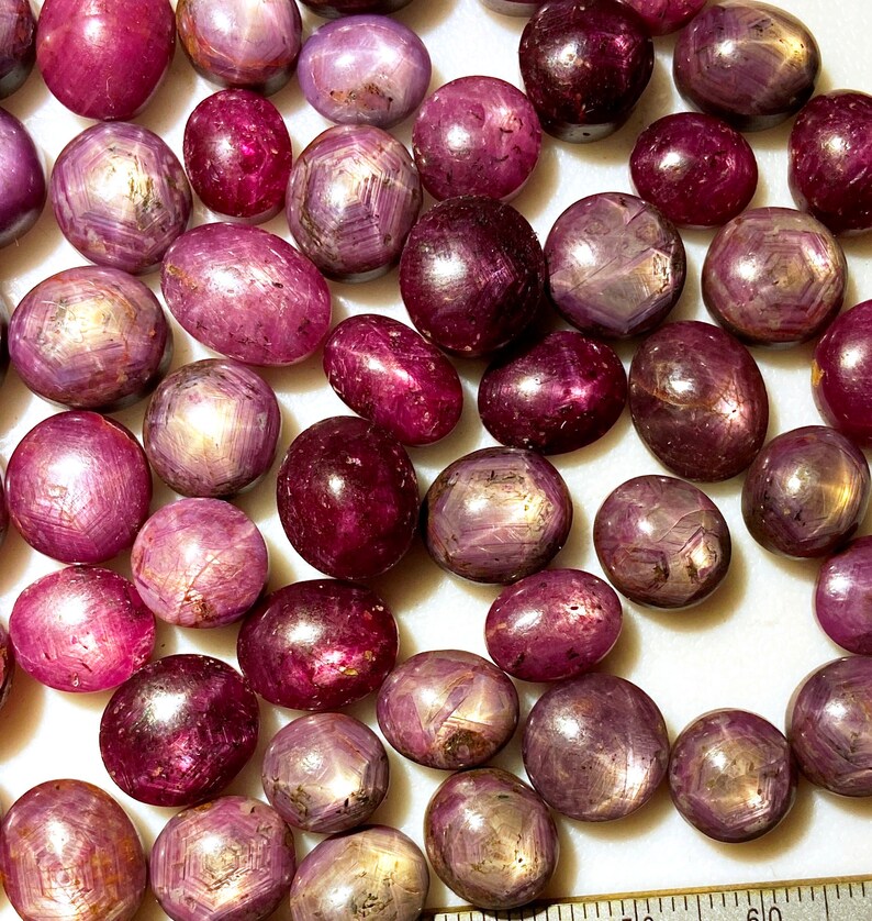 May include: A collection of pink star ruby cabochons, each with a six-pointed star in the center. The stones are various sizes and shapes, and they are all polished to a high shine.