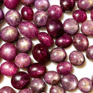 May include: A collection of loose, oval-shaped, pink and purple gemstones with a star-like pattern. The stones are arranged on a white surface.