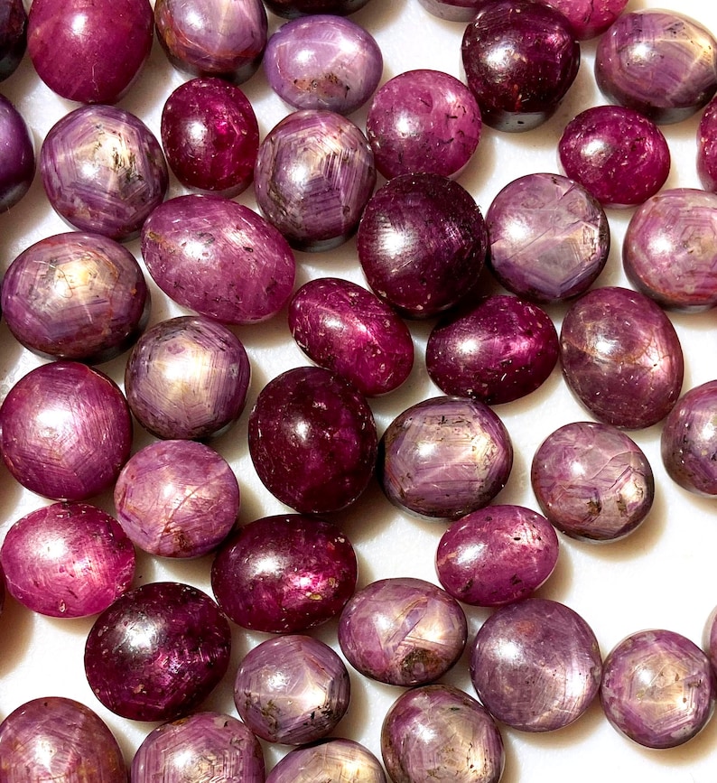 May include: A collection of loose, oval-shaped, pink and purple gemstones with a star-like pattern. The stones are arranged on a white surface.