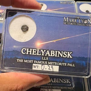 May include: A small, dark, textured Chelyabinsk meteorite fragment encased in a clear rectangular display case. The case features a blue graphic of a meteor streak, the text "THE MARK LYON METEORITE COLLECTION," "CHELYABINSK LL5," and "WT: 0.29."