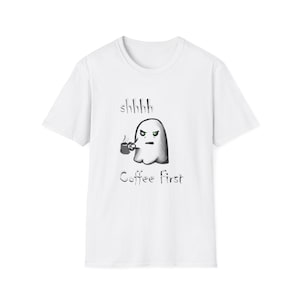 Puede incluir: Camiseta blanca con un fantasma gruñón sosteniendo una taza de café. El fantasma tiene ojos verdes y el texto "shhhhh Coffee First" está impreso encima y debajo de la ilustración del fantasma. La camiseta tiene mangas cortas y cuello redondo.