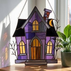 May include: A stained glass suncatcher shaped like a house with purple and black glass panels, yellow windows, a chimney, and a crescent moon. The piece features dark metal framing and is suspended by a silver chain for hanging in a window.