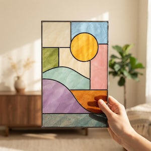 Pode incluir: Um painel retangular de vitral com uma composição geométrica de peças de vidro texturizado em tons de amarelo, azul, verde, rosa, roxo e laranja, com um círculo amarelo proeminente representando um sol em uma moldura de metal escuro.