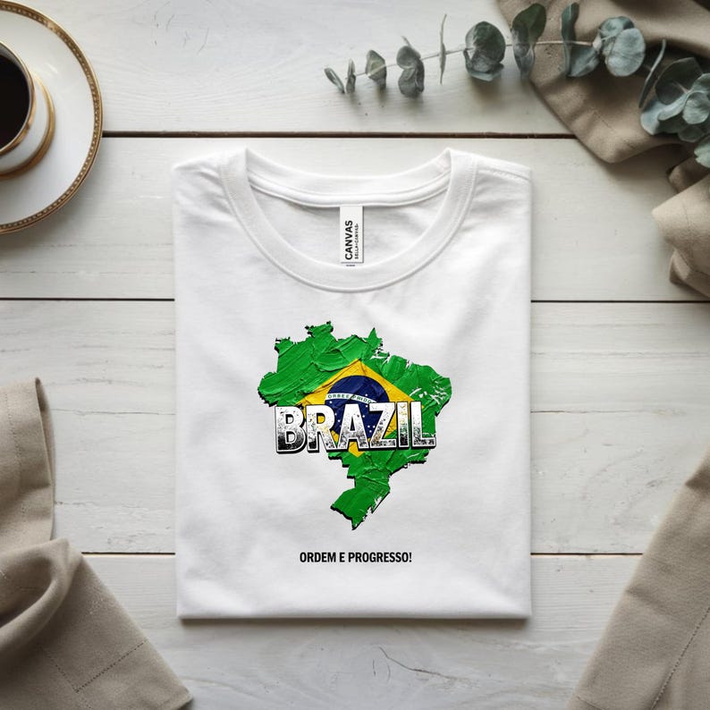 以下が含まれることがあります： ブラジルの緑と黄色の地図がプリントされた白いTシャツ。太字で縁取りされた「BRAZIL」の文字と「ORDEM E PROGRESSO！」のフレーズが特徴。Tシャツはきれいに折りたたまれています。愛国的なデザインです。