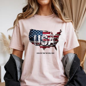 以下が含まれることがあります： アメリカ国旗のデザインが施された、アメリカ合衆国の地図が描かれたライトピンクのTシャツ。「USA」の文字は白でプリントされ、その下に「I BELIEVE THAT WE WILL WIN!」というテキストが入っています。