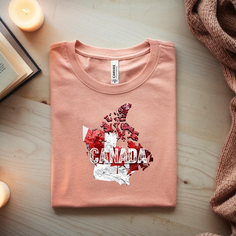 以下が含まれることがあります： カナダの国旗と白い「CANADA」の文字がプリントされた、ピーチ色のTシャツ。シャツはきれいに折りたたまれ、首元には「CANVAS」のブランドタグが見えます。デザインは赤と白のカナダの地図が特徴です。