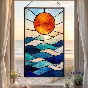 Könnte beinhalten: Ein rechteckiges Buntglas-Fensterbild mit einer leuchtend orangefarbenen Sonne über stilisierten Meereswellen in verschiedenen Blau-, Türkis- und Weißtönen. Das Stück ist in dunkles Metall gefasst und an einer dünnen Kette aufgehängt.