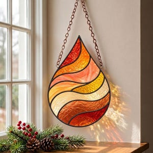 May include: A teardrop-shaped stained glass suncatcher featuring wavy horizontal panels in shades of red, orange, and yellow. The glass piece is framed in dark metal and suspended by a matching copper-toned metal chain for hanging display.