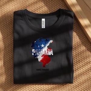 以下が含まれることがあります： パナマの地図が描かれた黒いTシャツ。パナマ国旗の色である青、白、赤でデザインされています。「PANAMA」の文字は白でプリントされ、その下に「VAMOS PANAMA！」と書かれています。Tシャツは、質感のある表面に折りたたまれています。