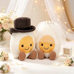 May include: Two plush egg-shaped toys with white bodies and yellow centers. One wears a dark brown top hat, and the other wears a white tulle veil. Both have small black eyes, stitched smiles, and brown corduroy feet. A small white ring box sits nearby.